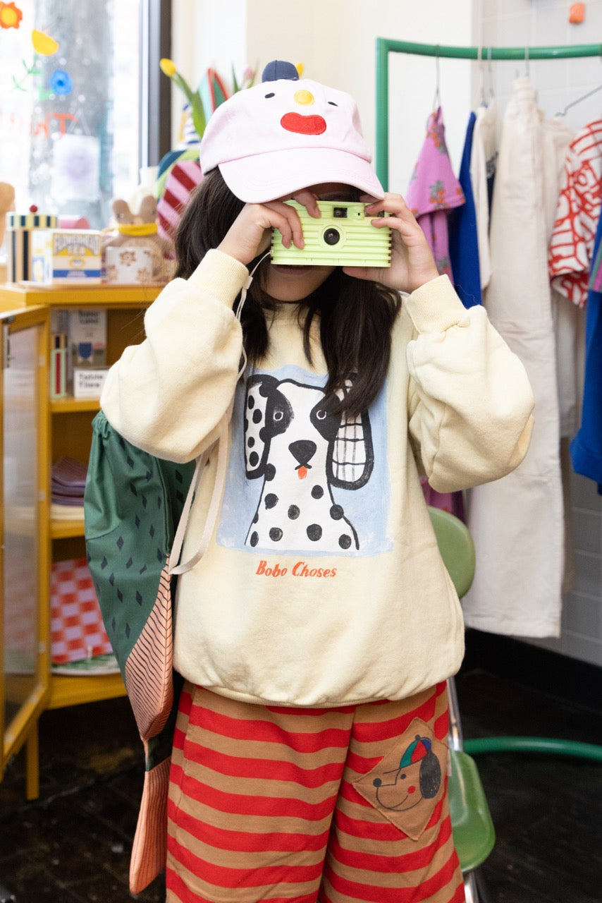 A child wearing a cream-colored sweatshirt with a Dalmatian graphic and the text 'Bobo Choses' under it, paired with red and beige striped pants and a light green striped camera. The child is holding a retro-style green camera to their face in a retail store setting.